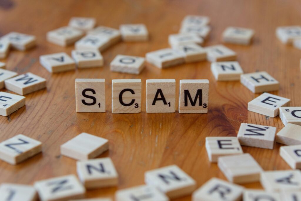 Wooden letter tiles spelling “SCAM” on a table, representing fraud, financial scams and online security risks.